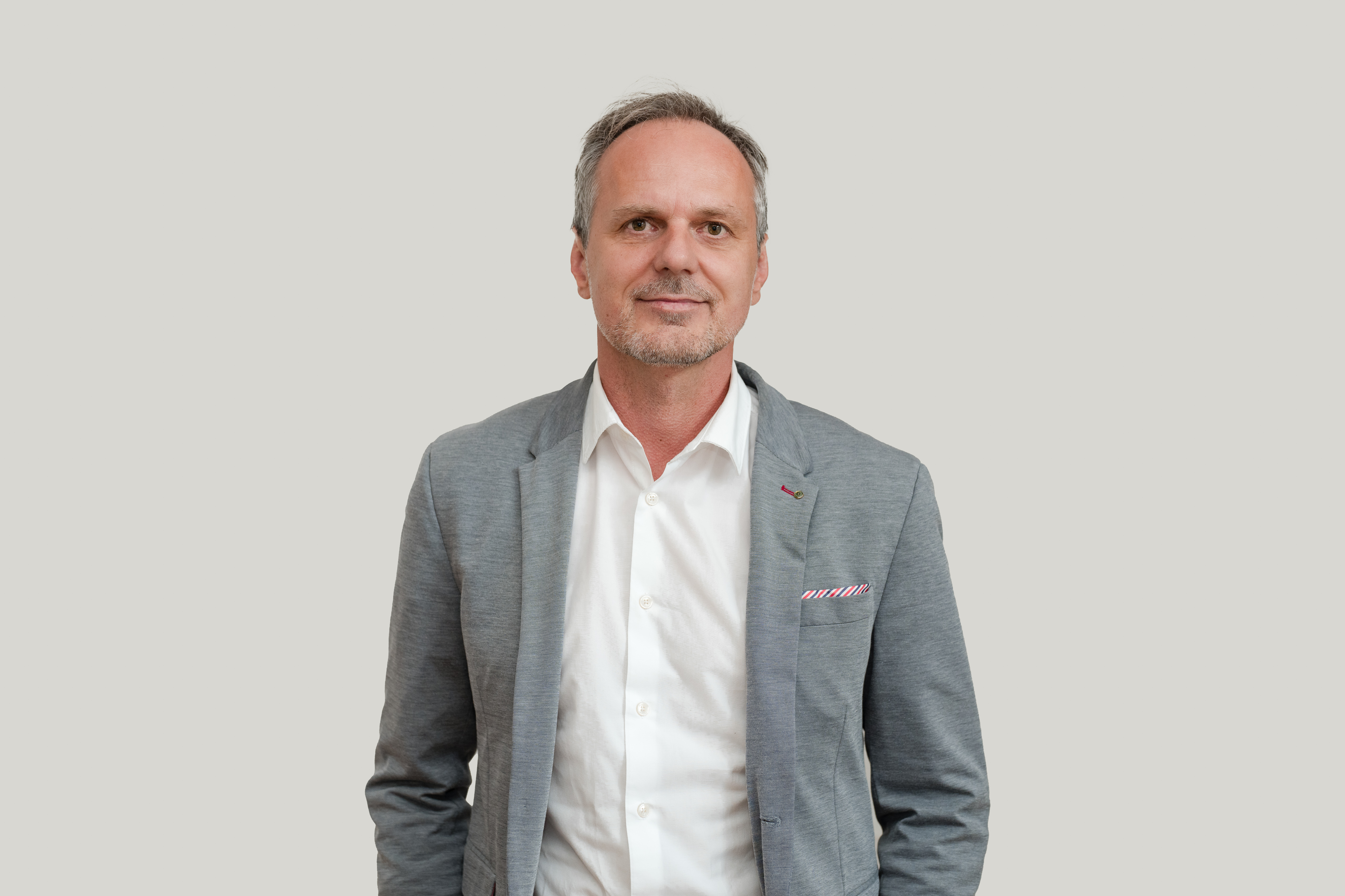 Mittelalter Mann im grauen Anzug und weißem Hemd vor grauem Hintergrund, steht freundlich lächelnd.