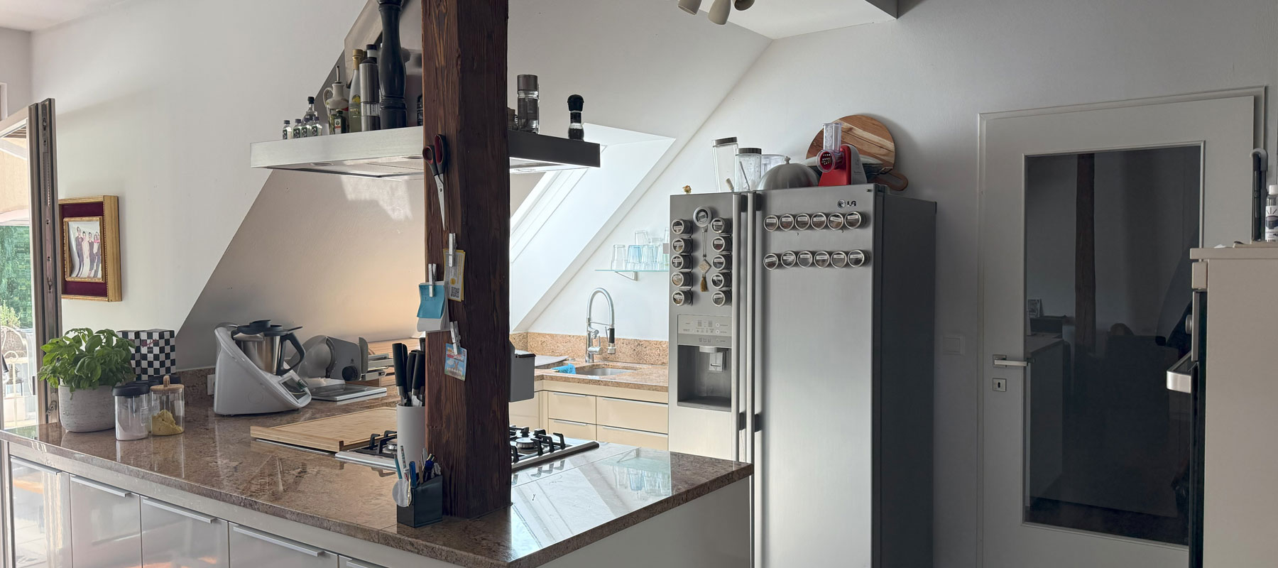 Moderne K&uuml;che mit Ger&auml;ten, Gew&uuml;rzdosen am K&uuml;hlschrank, Holzs&auml;ule, Pflanzen, Bild an der Wand.